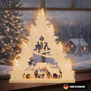 Op de afbeelding: Houten kerstversiering in de vorm van een gestileerde bosscène. De scène omvat herten, bomen en een kleine hut, allemaal verlicht door warme lichten. De decoratie is geplaatst tegen een besneeuwde winterachtergrond. De woorden "MADE IN GERMANY" zijn zichtbaar.