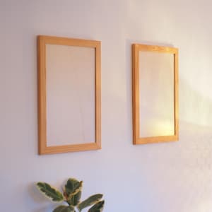 Puede incluir: Dos marcos de fotos rectangulares de madera clara están montados en una pared blanca. Los marcos tienen un diseño sencillo y el cristal es transparente. Una planta verde con hojas abigarradas es visible en la parte inferior.