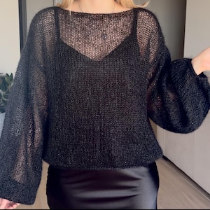 Può includere: Maglione nero a maglia con scollatura ampia e maniche lunghe e voluminose. Il maglione è semitrasparente, rivelando una canotta nera sotto. Il fondo del maglione è corto e le maniche hanno un polsino arricciato. Il maglione è abbinato a una gonna di raso nera.