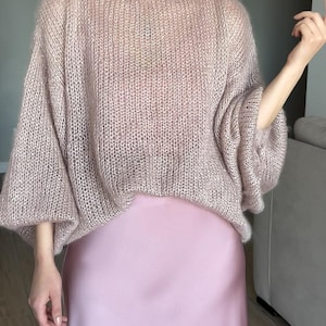 Può includere: Un maglione lavorato a maglia rosa chiaro oversize con scollo a barchetta e maniche lunghe e voluminose. Il maglione è abbinato a una gonna di raso rosa chiaro. La maglieria ha un aspetto morbido e strutturato e lo stile generale è casual e confortevole.