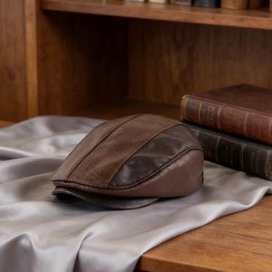 May include: A brown leather flat cap with a dark brown brim and a textured surface. The cap is resting on a silver satin cloth, with two leather-bound books in the background.