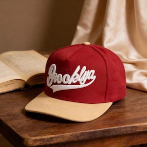 May include: A burgundy baseball cap with a tan brim. The word "Brooklyn" is embroidered in white script across the front. The cap is sitting on a wooden surface next to an open book.