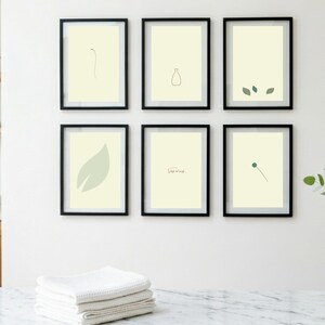 Może przedstawiać: Sześć oprawionych wydruków artystycznych z minimalistycznymi botanicznymi rysunkami liniowymi w czarnych ramkach. Wydruki przedstawiają proste ilustracje liści, łodyg i wazonu na kremowym tle. Stos białych ręczników znajduje się na pierwszym planie.