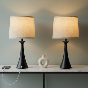 May include: Two black table lamps with beige fabric shades sit on a white marble-topped table. A small white vase and a smartphone charging with a white cable are also on the table. The lamps have a classic, elegant design.