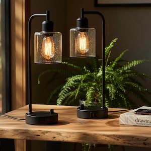 May include: Two black metal table lamps with clear glass shades and exposed filament bulbs. The lamps sit on a wooden table with a book and a phone. A fern is in the background.