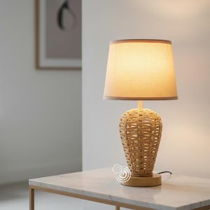 May include: A beige table lamp with a woven rattan base and a beige lampshade. The lamp sits on a marble-topped table. The lamp is turned on, illuminating the room.