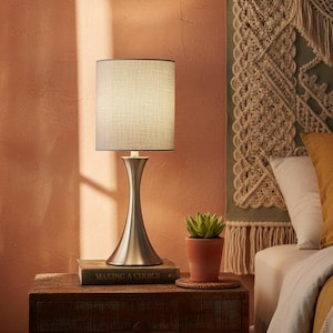 May include: A brushed nickel table lamp with a linen shade sits on a wooden nightstand. A small potted succulent and a book titled "MAKING A CHOICE" are also on the nightstand. The background features a textured wall and a decorative wall hanging.