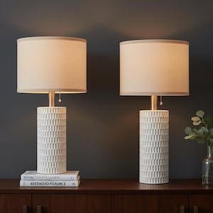 May include: Two matching table lamps with cream-colored shades and textured white ceramic bases. The lamps sit on a dark wood surface, with a small vase of greenery to the right. The lamps are lit, casting a warm glow.
