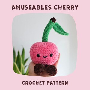 May include: A pink crocheted cherry plush toy with a smiling face, green stem, and brown feet. The text "AMUSEABLES CHERRY" and "CROCHET PATTERN" are displayed above and below the toy, respectively. The background is a light pink color.