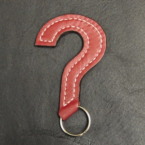 May include: A red leather question mark keyring with white stitching. The keyring has a silver ring.