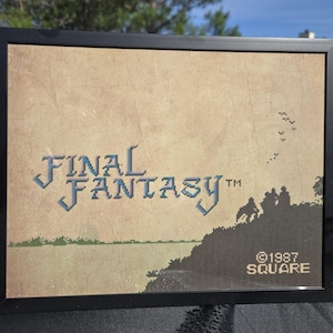 May include: A framed print featuring the pixelated title Final Fantasy in blue text against a tan background. The image includes a dark silhouette of figures on a hill, a small flock of birds, and the text 1987 SQUARE in the bottom right corner.