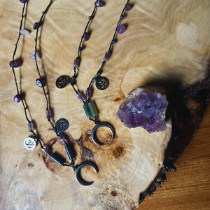 May include: Two necklaces with black cords, amethyst beads, and silver crescent moon pendants. One necklace has an Om symbol pendant. A raw amethyst crystal sits on a wooden surface.