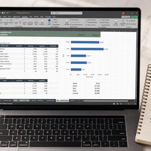 May include: A laptop screen displaying a spreadsheet titled API & REPORTING - PLUMBING, featuring tables and a horizontal bar chart that visualises job counts and revenue data by service type and zone, with a dark grey keyboard and a black bezel.