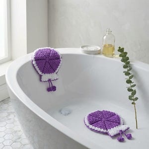 Puede incluir: Dos esponjas de baño de ganchillo moradas y blancas hechas a mano en un baño. Una esponja está en posición vertical y la otra está plana. Las esponjas tienen un diseño circular con un borde festoneado y una borla decorativa.