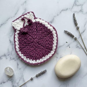 Può includere: Un portasapone fatto a mano, all'uncinetto, di colore viola intenso con un bordo bianco e chiusura con coulisse. Il portasapone è accanto a una saponetta e rametti di lavanda.