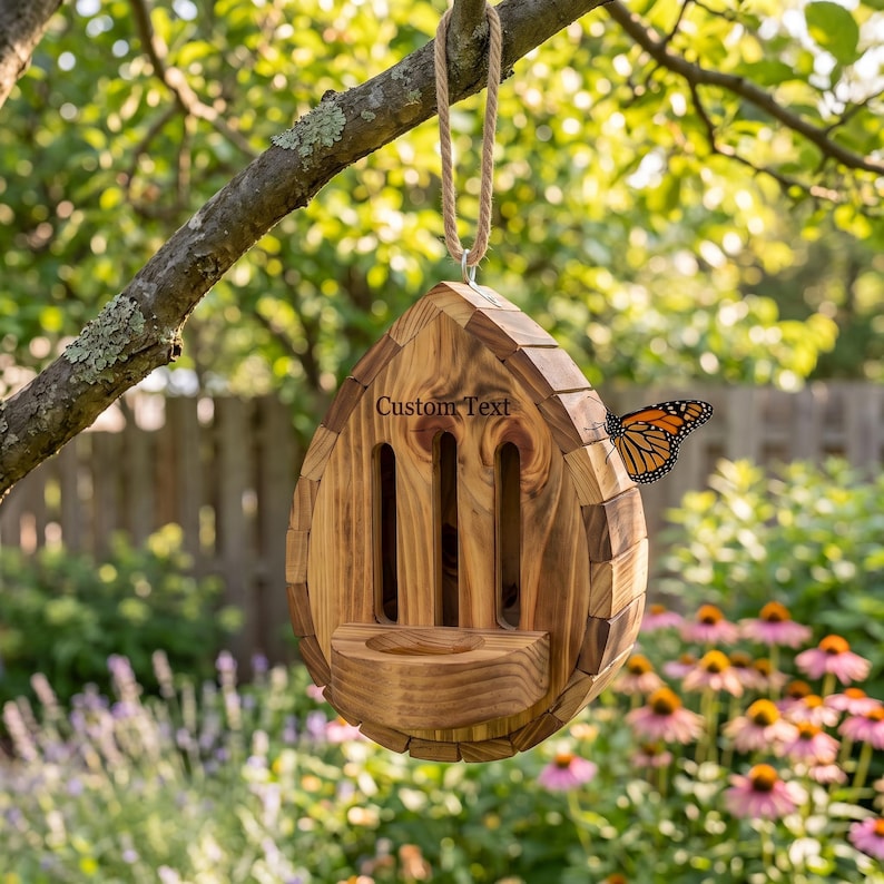 May include: A teardrop-shaped wooden butterfly house with a light brown wood grain finish, featuring three vertical entry slots, a small landing platform, and a rope hanger. The text Custom Text is engraved on the front, with a monarch butterfly.