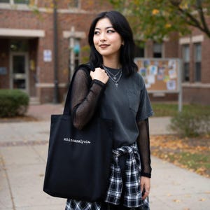 Puede incluir: Bolso tote negro con la palabra "schizoanalysis." impresa en blanco. El bolso se lleva sobre el hombro. La persona lleva una camiseta negra, una camiseta de manga larga de malla negra y una camisa a cuadros en blanco y negro atada a la cintura.