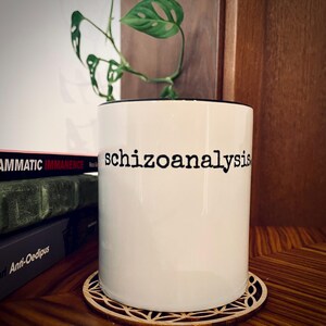 Puede incluir: Una taza de cerámica blanca con la palabra "schizoanalysis" impresa en negro. La taza está sobre un posavasos de madera con un diseño geométrico. Una planta verde con hojas únicas está en el fondo, junto con una pila de libros.