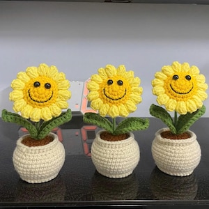 Puede incluir: Tres girasoles de ganchillo con caras sonrientes en macetas color crema. Los girasoles tienen pétalos amarillos, ojos negros y hojas verdes. Las macetas están llenas de hilo marrón, que simula tierra. Un artículo decorativo alegre.