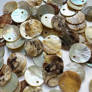 Può includere: Una collezione di perline rotonde e iridescenti a forma di conchiglia in vari colori, tra cui crema, marrone e oro. Ogni perlina ha un piccolo foro per l'infilatura. Le perline hanno un aspetto naturale e strutturato, adatte per la creazione di gioielli o lavori artigianali.