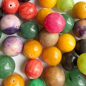 Op de afbeelding: Een verzameling kleurrijke, ronde kralen in verschillende tinten rood, roze, geel, groen en paars. De kralen hebben een glad, gepolijst oppervlak en zijn waarschijnlijk gemaakt van steen of glas. Elke kraal heeft een klein gaatje.