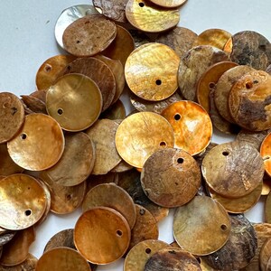 Könnte beinhalten: Eine Sammlung runder, flacher Muschelkernperlen in Braun-, Gold- und Beigetönen. Jede Perle hat ein kleines Loch in der Nähe der Oberseite. Die Perlen variieren in Farbe und Textur, wobei einige glatt und andere strukturierter erscheinen.