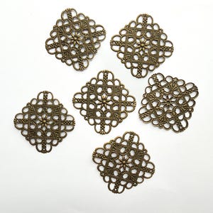 Puede incluir: Seis componentes de filigrana de metal de forma cuadrada con un acabado en bronce antiguo. Cada pieza presenta un patrón calado simétrico e intrincado con un motivo central en forma de estrella y bordes delicados con bucles.