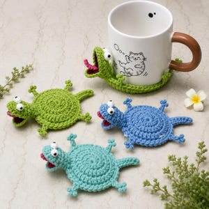 May include: A white ceramic mug with a brown handle rests on a green crocheted lizard coaster. Three additional crocheted lizard coasters in green, blue, and teal are scattered around. Each lizard has googly eyes and a pink tongue.