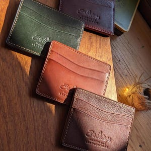 May include: A collection of handcrafted leather cardholders in various colors, including green, brown, and burgundy. Each wallet features a minimalist design with visible stitching and the brand's logo. A vintage tobacco tin is in the background.