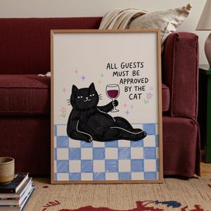 May include: A framed wall art print featuring a black cat lounging on a blue and white checkered floor, holding a glass of red wine. The text reads, ALL GUESTS MUST BE APPROVED BY THE CAT, accompanied by small colorful star and flower illustrations.