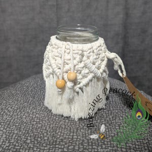May include: A clear glass jar with a cream-colored macrame cozy. The cozy has a woven pattern with wooden beads and fringe. The jar is on a gray patterned surface. The words "Buzzing Peacock" are visible.