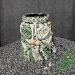 May include: A handmade macrame water bottle holder in shades of green and white. The holder features a braided top, a drawstring with wooden beads, and a fringe at the bottom. A brown tag is attached to the side.