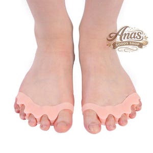 May include: A pair of feet wearing peach-colored toe separators. The separators are made of a soft, flexible material and are designed to fit between the toes. The image includes the text "Anas GOODS SHOP".