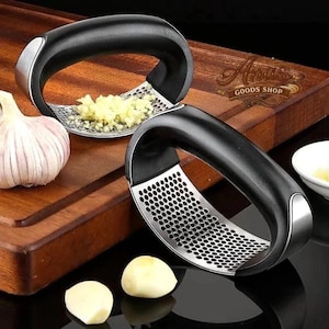 May include: Two black and silver garlic presses with minced garlic. A whole garlic bulb and two cloves are also visible. The presses have a curved design with a perforated pressing surface.
