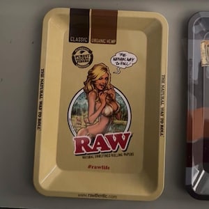Pu&ograve; includere: Un vassoio rettangolare giallo chiaro con un bordo marrone. Il vassoio presenta una grafica di una donna e il testo "RAW" in rosso. Il testo aggiuntivo include "Classic Organic Hemp", "The Natural Way to Roll" e "Natural Unrefined Rolling Papers."