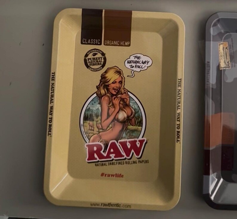Pu&ograve; includere: Un vassoio rettangolare giallo chiaro con un bordo marrone. Il vassoio presenta una grafica di una donna e il testo "RAW" in rosso. Il testo aggiuntivo include "Classic Organic Hemp", "The Natural Way to Roll" e "Natural Unrefined Rolling Papers."