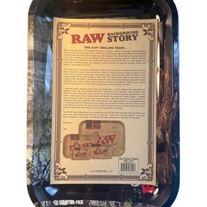 May include: A rectangular black rolling tray with a detailed paper insert. The insert features the text "RAW" and "BACKGROUND STORY" with a vintage aesthetic. The tray is designed for rolling tobacco and has a forest-themed border.