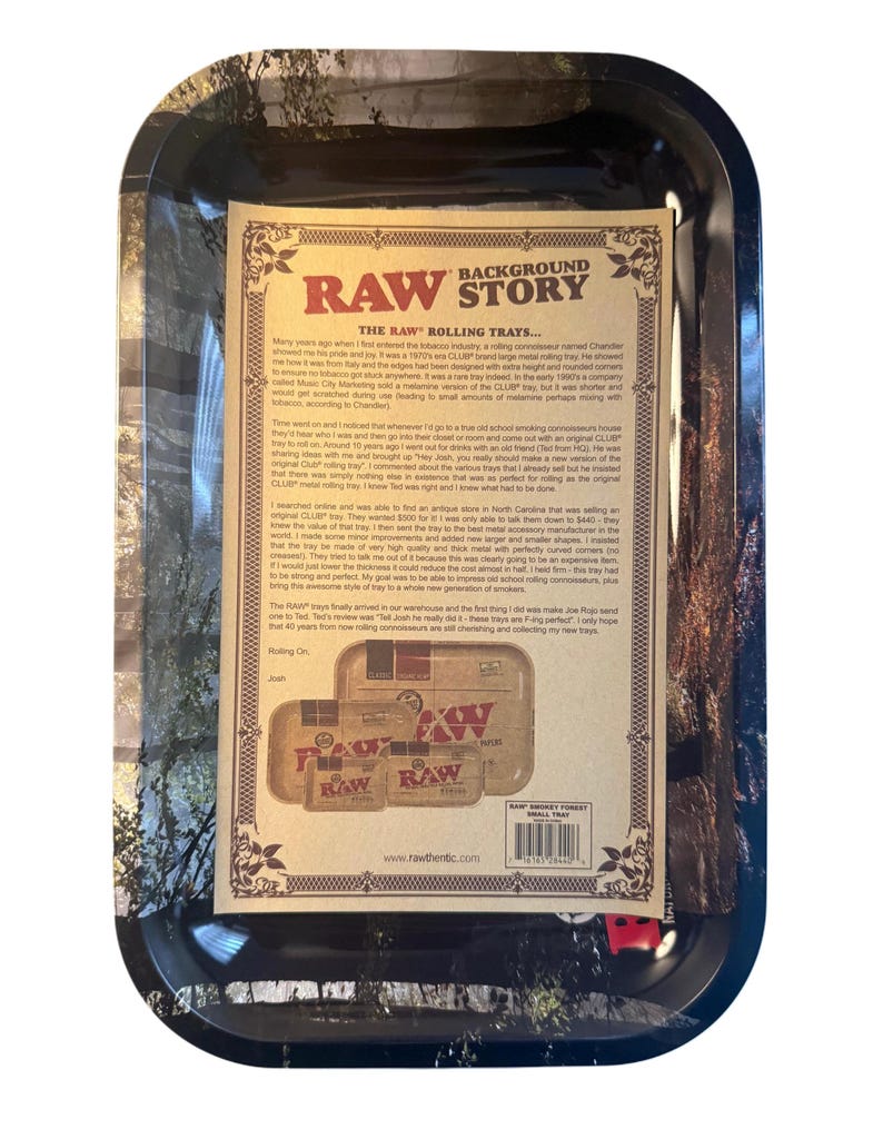 May include: A rectangular black rolling tray with a detailed paper insert. The insert features the text "RAW" and "BACKGROUND STORY" with a vintage aesthetic. The tray is designed for rolling tobacco and has a forest-themed border.
