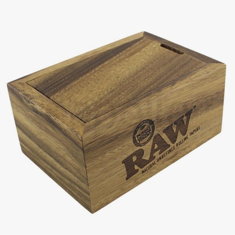 K&ouml;nnte beinhalten: Rechteckige Holzbox mit Schiebedeckel aus hellbraunem Holz. Auf der Vorderseite befindet sich das "RAW"-Logo und der Text. F&uuml;r nat&uuml;rliche, unraffinierte Zigarettenpapiere.