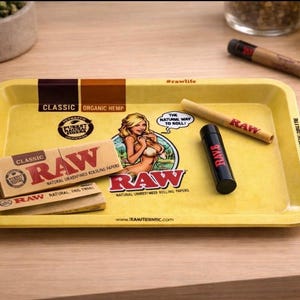 Pu&ograve; includere: Vassoio giallo per rollare con marchio RAW, un'immagine a cartoni animati e testo. Include un pacchetto di cartine, un libretto di cartine, un cono pre-rollato e un contenitore nero. Il vassoio ha il testo "CLASSIC ORGANIC HEMP".