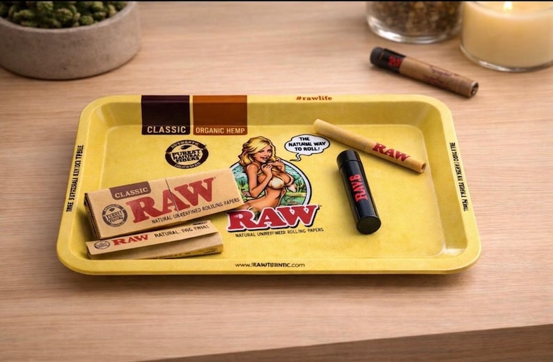 Pu&ograve; includere: Vassoio giallo per rollare con marchio RAW, un'immagine a cartoni animati e testo. Include un pacchetto di cartine, un libretto di cartine, un cono pre-rollato e un contenitore nero. Il vassoio ha il testo "CLASSIC ORGANIC HEMP".