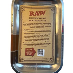 May include: A silver metal rolling tray with rounded corners, holding a brown certificate of authenticity. The certificate has black text and a QR code. The tray features the text "RAW" and "THE NATURAL WAY TO ROLL".