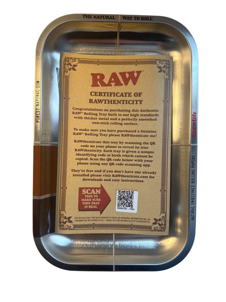 May include: A silver metal rolling tray with rounded corners, holding a brown certificate of authenticity. The certificate has black text and a QR code. The tray features the text "RAW" and "THE NATURAL WAY TO ROLL".