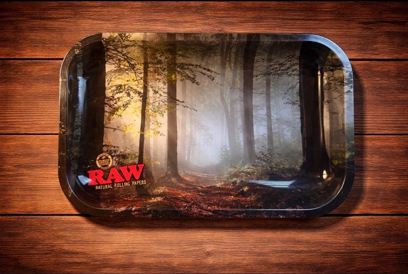 May include: Rectangular metal rolling tray with rounded corners. The tray features a forest scene with a path leading into the distance. The RAW logo is in the lower left corner. The tray is on a wooden surface.