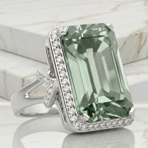 May include: A silver ring with a large, rectangular, pale green gemstone. The gemstone is framed by small, clear stones. The ring has a split shank design with additional small stones on the side.