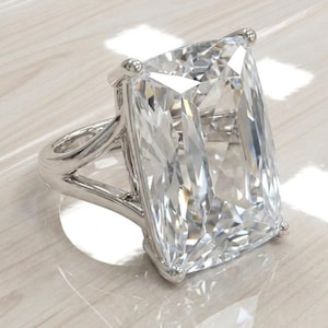 May include: A large, rectangular-cut diamond ring with a silver band. The clear, faceted diamond reflects light. The ring has a split shank design. The ring is displayed on a light-coloured surface.