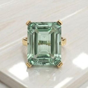 Puede incluir: Un anillo de oro con una gran piedra preciosa de color verde claro de corte esmeralda rectangular. La piedra está sujeta por cuatro garras de oro en cada esquina, mostrando una superficie facetada clara que refleja la luz sobre su estructura geométrica.