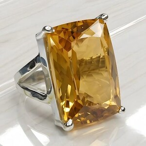 Puede incluir: Anillo de plata con una gran gema de citrino rectangular. La gema es de un color amarillo dorado vibrante y está sujeta por cuatro garras. El anillo tiene un diseño moderno y geométrico.