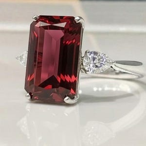 May include: A silver ring with a large, rectangular, deep red gemstone. Two smaller, pear-shaped, clear gemstones are set either side of the main stone. The ring's band is smooth and polished, reflecting light.