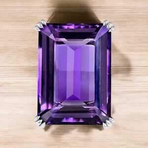 May include: A large, rectangular amethyst gemstone in a deep purple shade. The gemstone features a faceted, geometric design and is secured by four silver-coloured prongs. The stone is set against a light wood grain background.